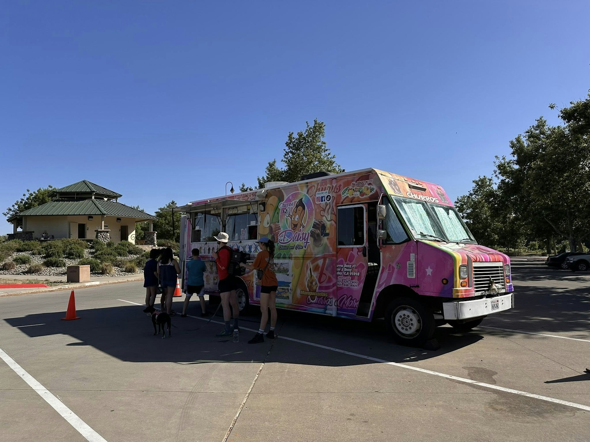A pink food truck serving customers in a parking lot, with a small building and trees in the background.