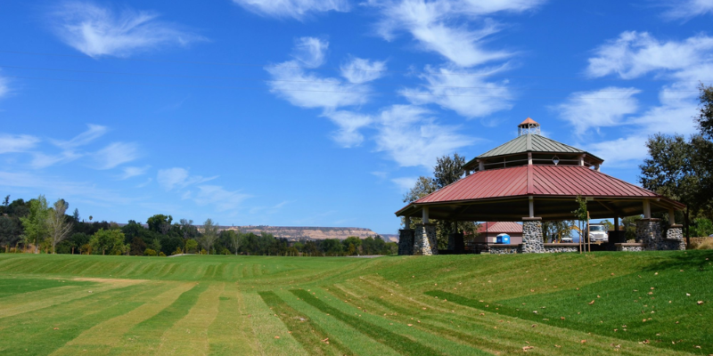 A gazebo with a red roof in a green park with striped lawn under a blue sky with wispy clouds.