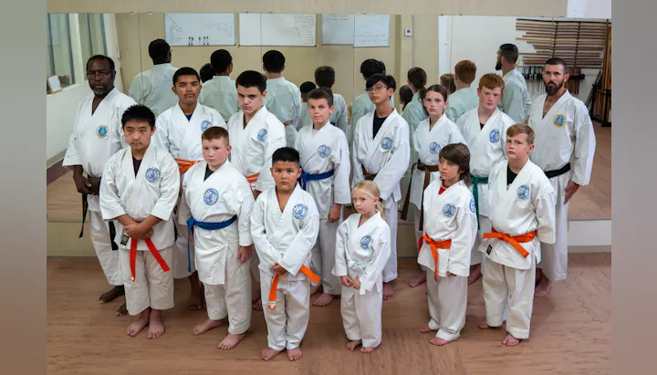 A group of children and adults in karate uniforms pose together. Some wear colored belts, indicating different skill levels.