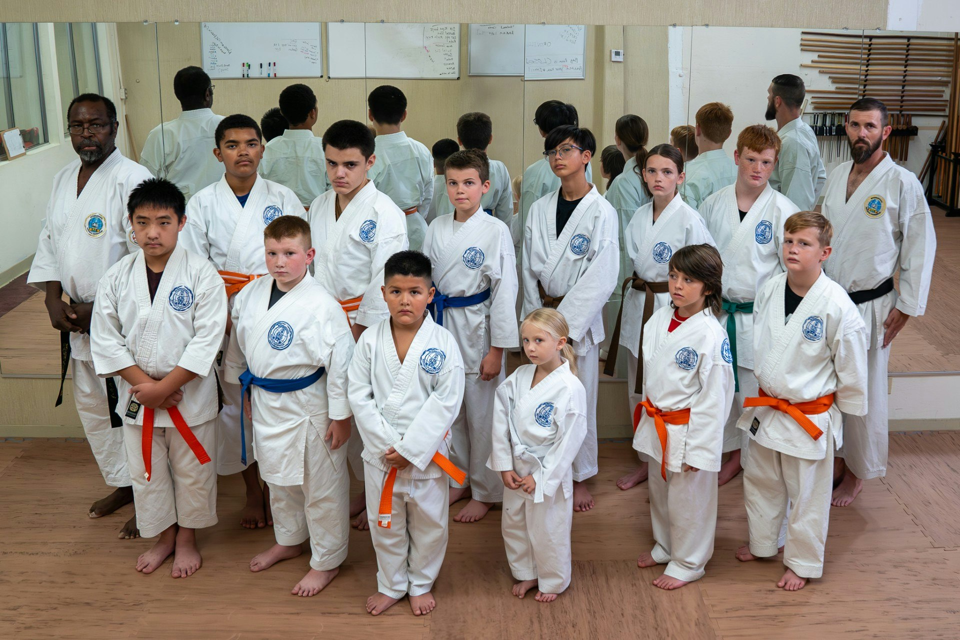 A group of children and adults in karate uniforms pose together. Some wear colored belts, indicating different skill levels.