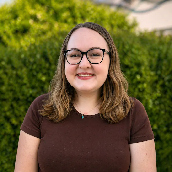 A smiling individual with glasses and shoulder-length hair stands in front of a green background, wearing a brown shirt.
