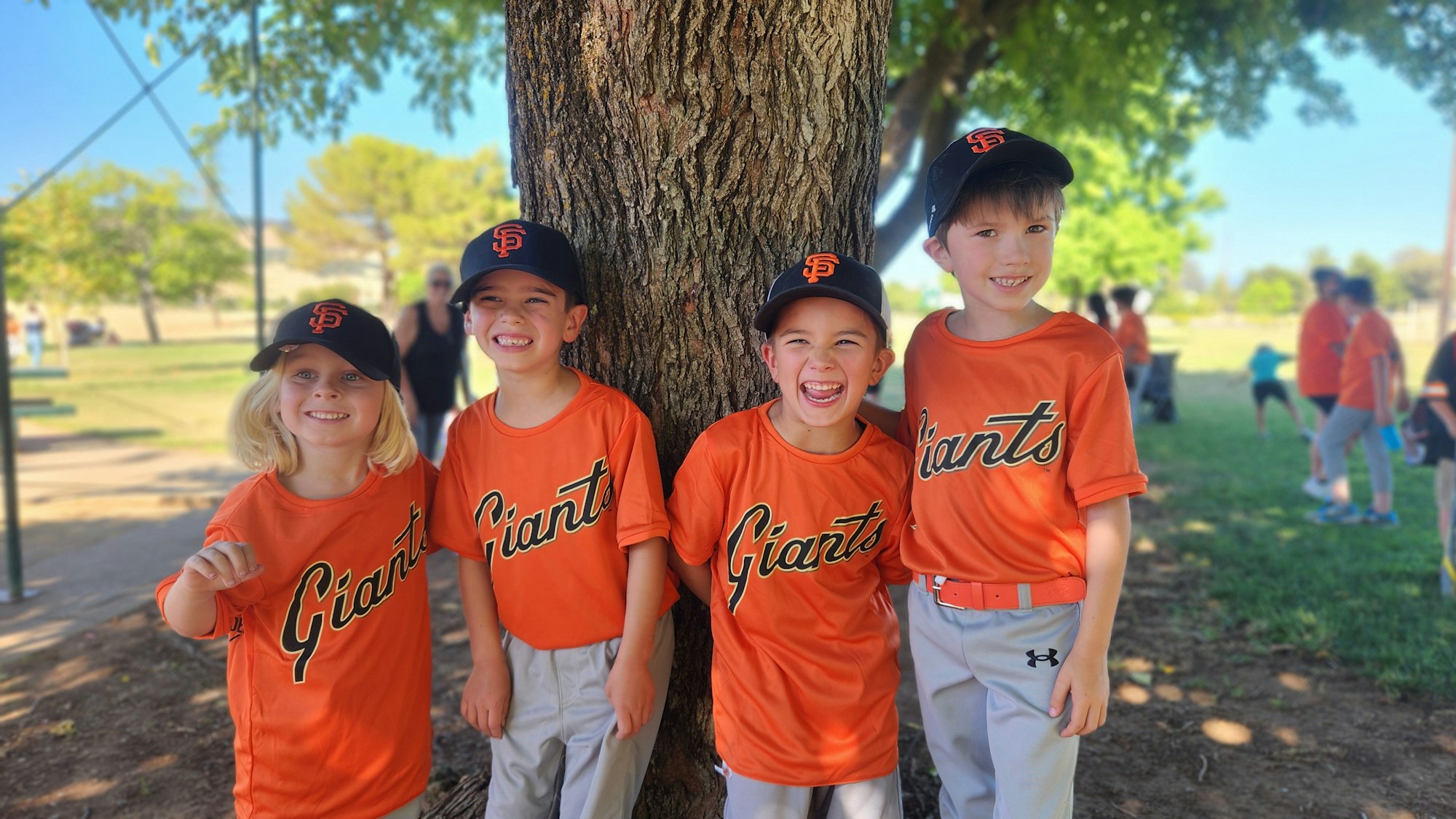 Four kids in orange "Giants" jerseys and black caps stand by a tree, smiling.