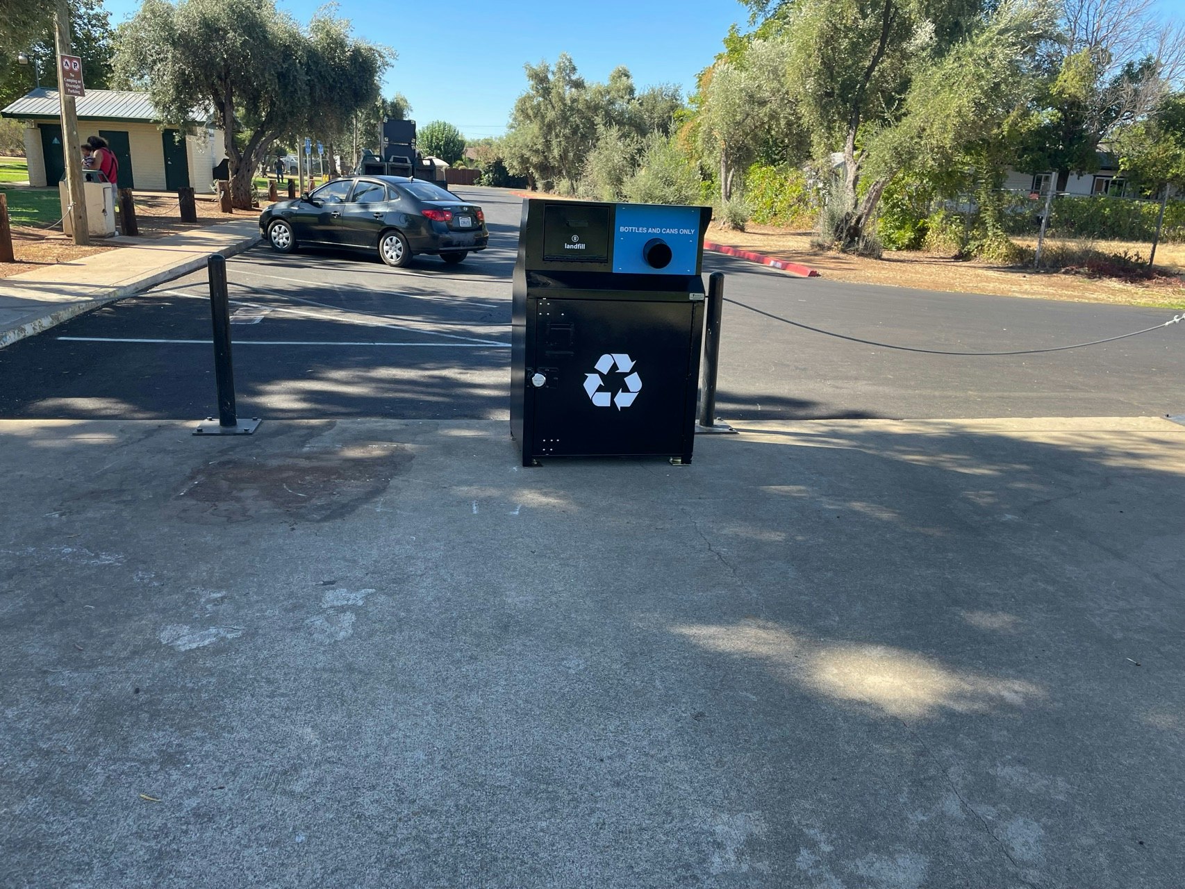 A recycling bin with a detached cable barrier in a parking area, trees in the background, and a person by a car.