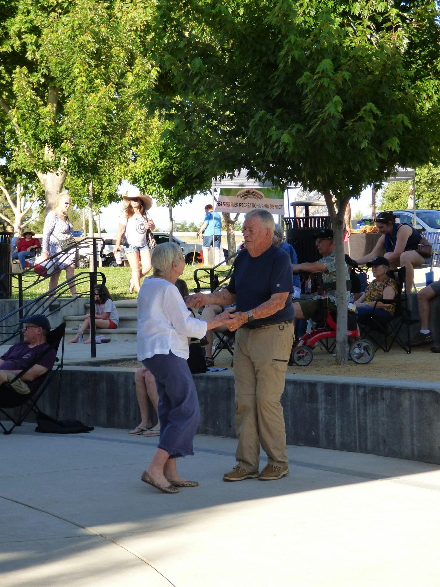 An older couple dances together in an outdoor park setting with people relaxing and enjoying the day.