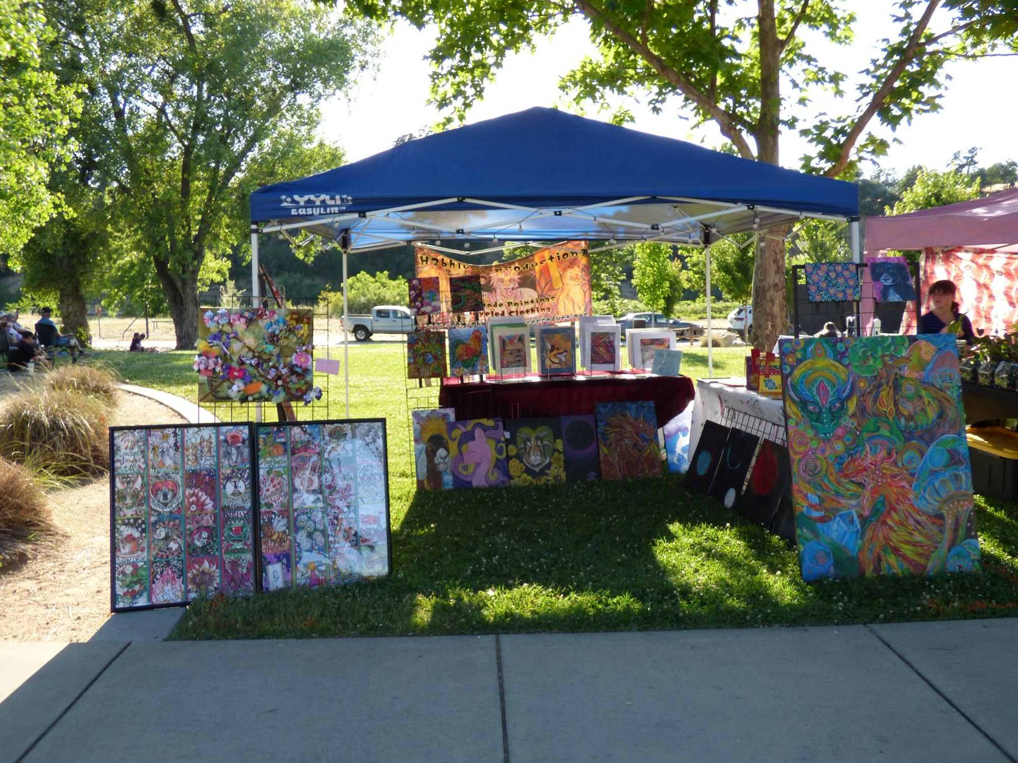 Outdoor art booth with colorful paintings and crafts displayed under a blue canopy, surrounded by green trees and grassy area.