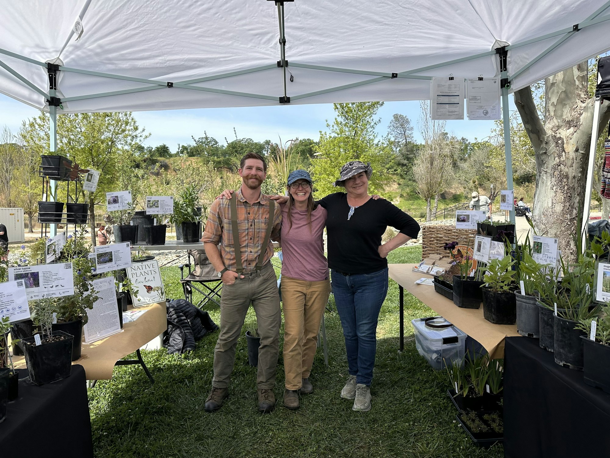 Three people smiling under a canopy at a plant stand with various potted plants and informational signs.