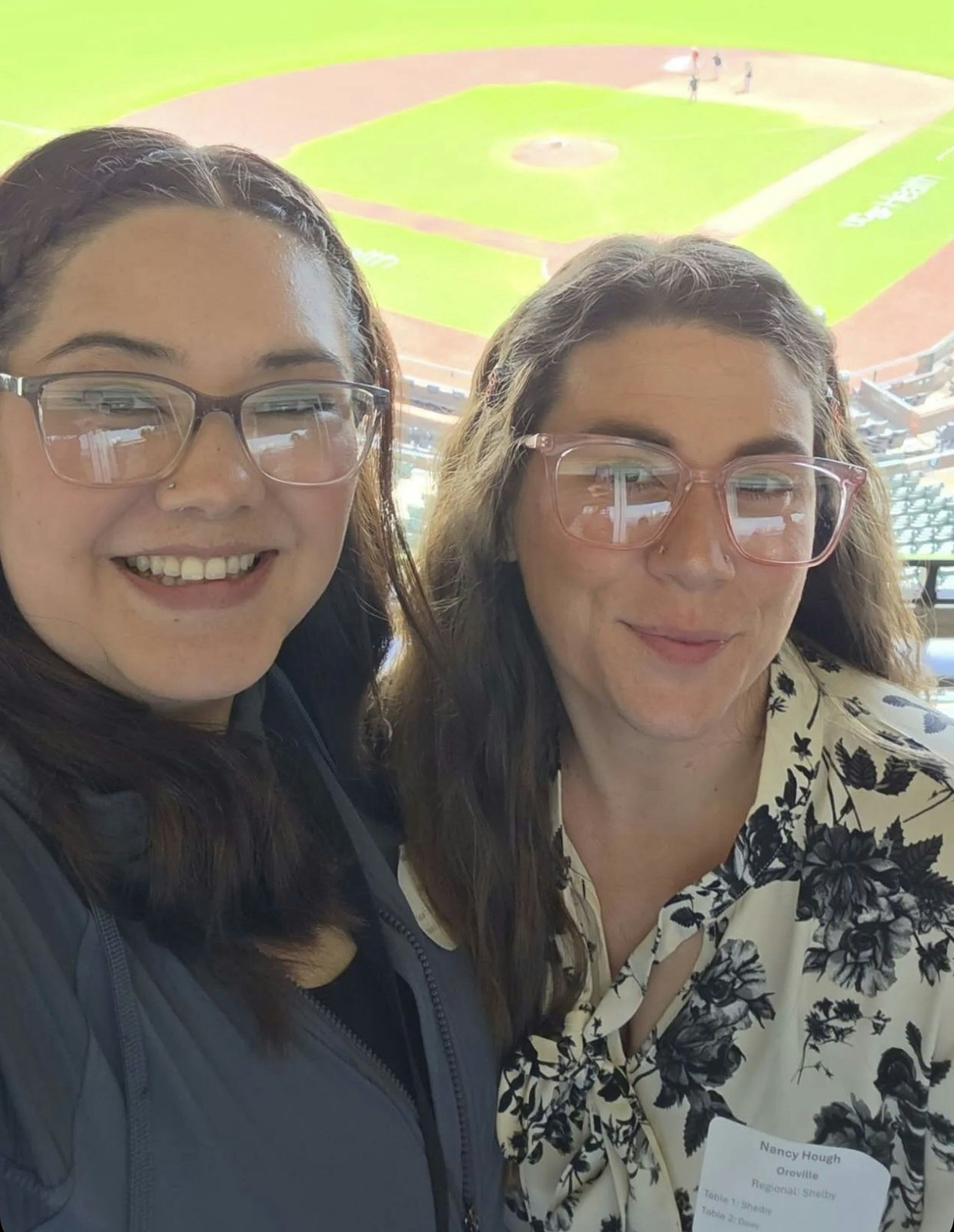Two women smiling for a selfie at a baseball venue, with a field visible in the background.