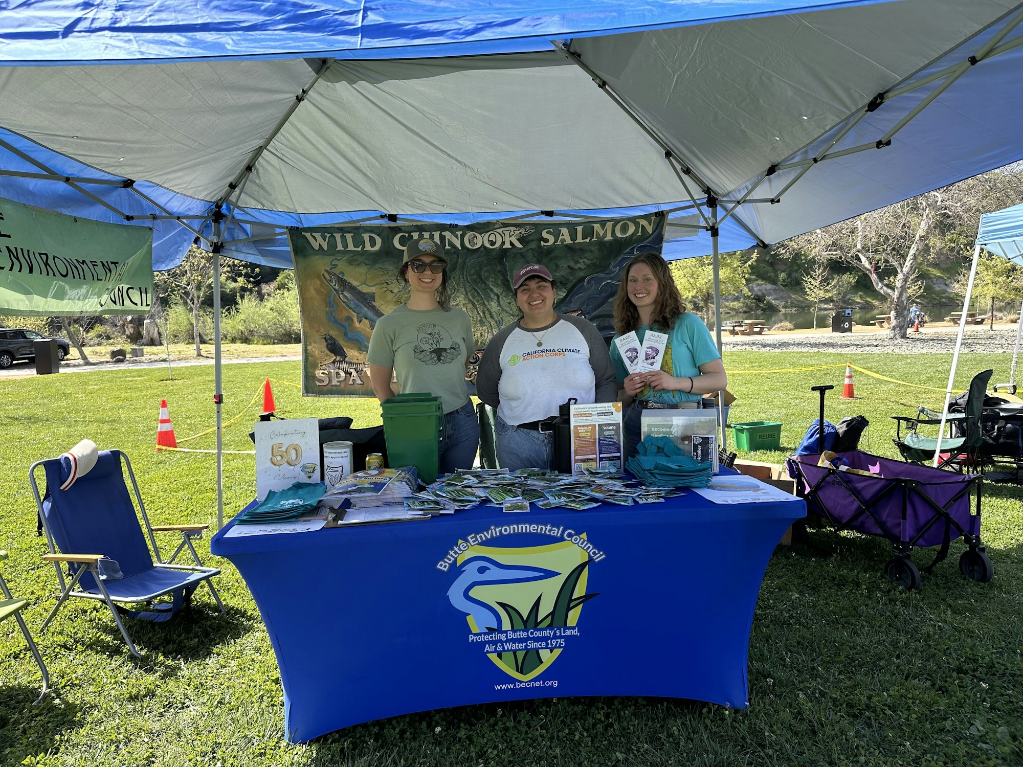 Outdoor booth with three people, brochures, and a banner about Wild Chinook Salmon, under a blue canopy.