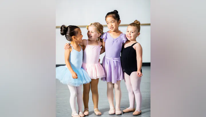 Four young girls in colorful ballet outfits, standing together, smiling and embracing each other.