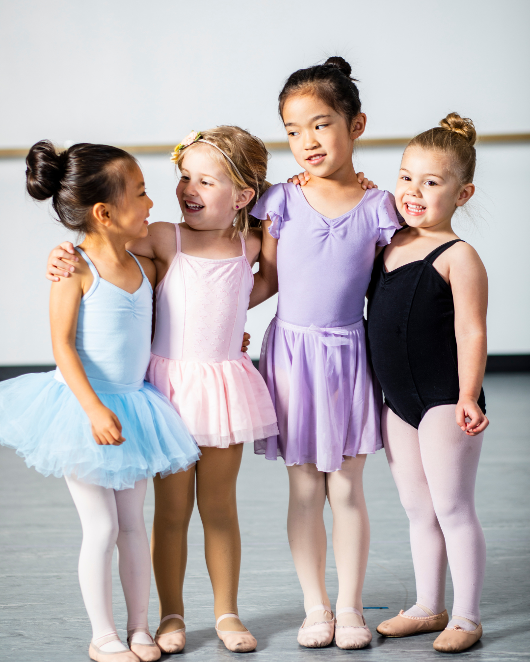 Four young girls in colorful ballet outfits, standing together, smiling and embracing each other.