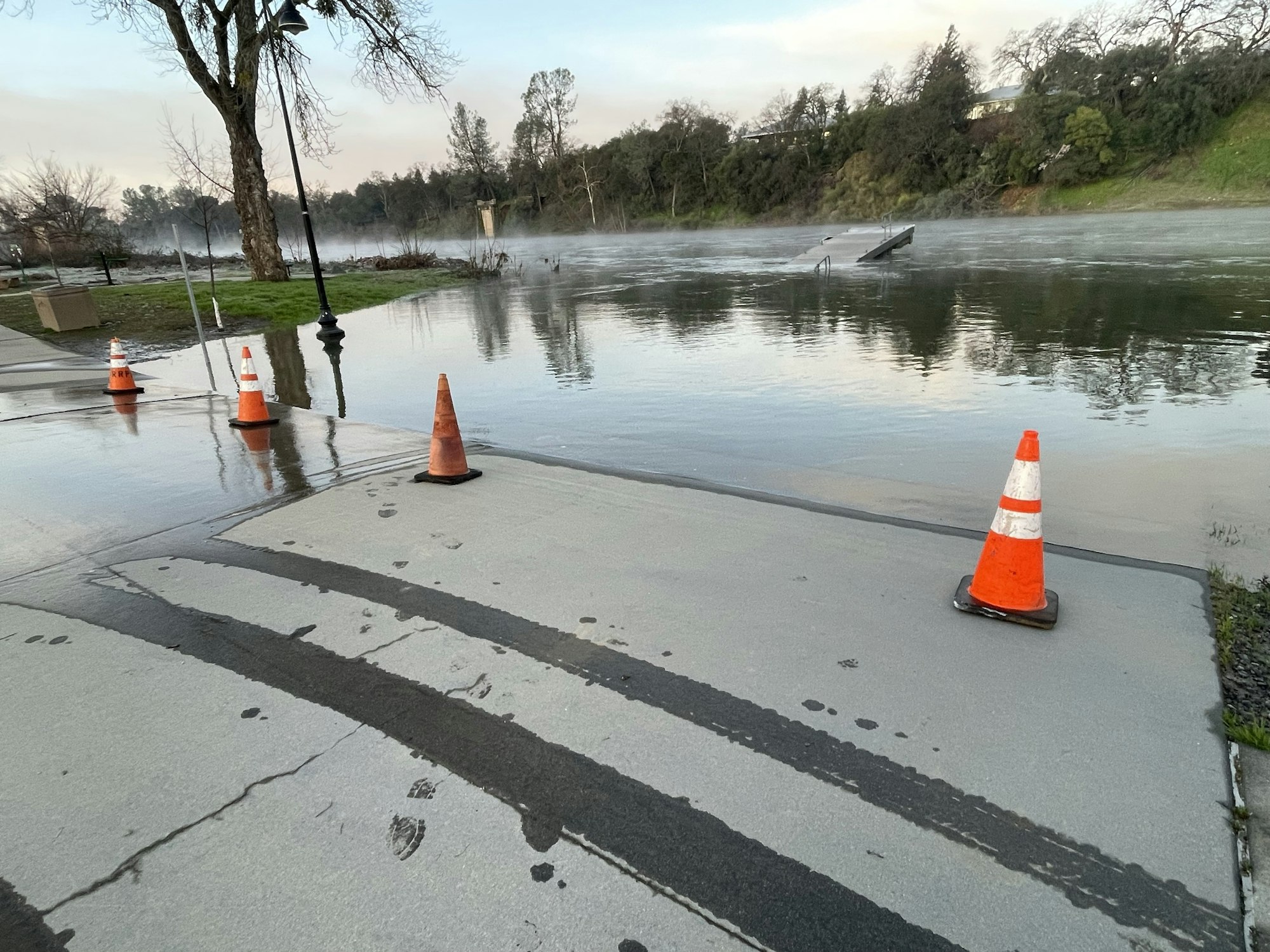 Flooded walkway with traffic cones by a river, mist in the background.