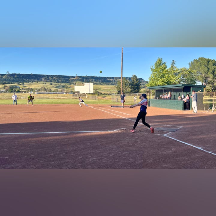 A softball game with a batter at the plate, fielders positioned, and spectators watching from the side.