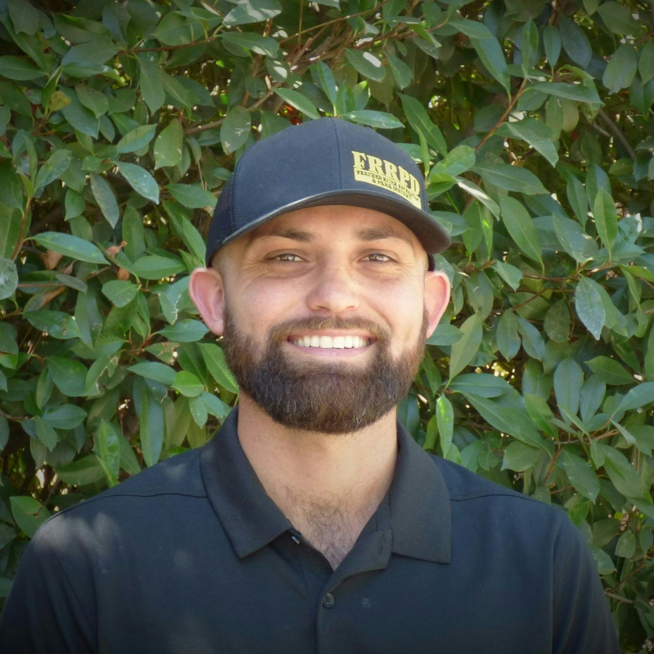 A smiling man with a beard, wearing a black cap and shirt, stands in front of a green leafy background.