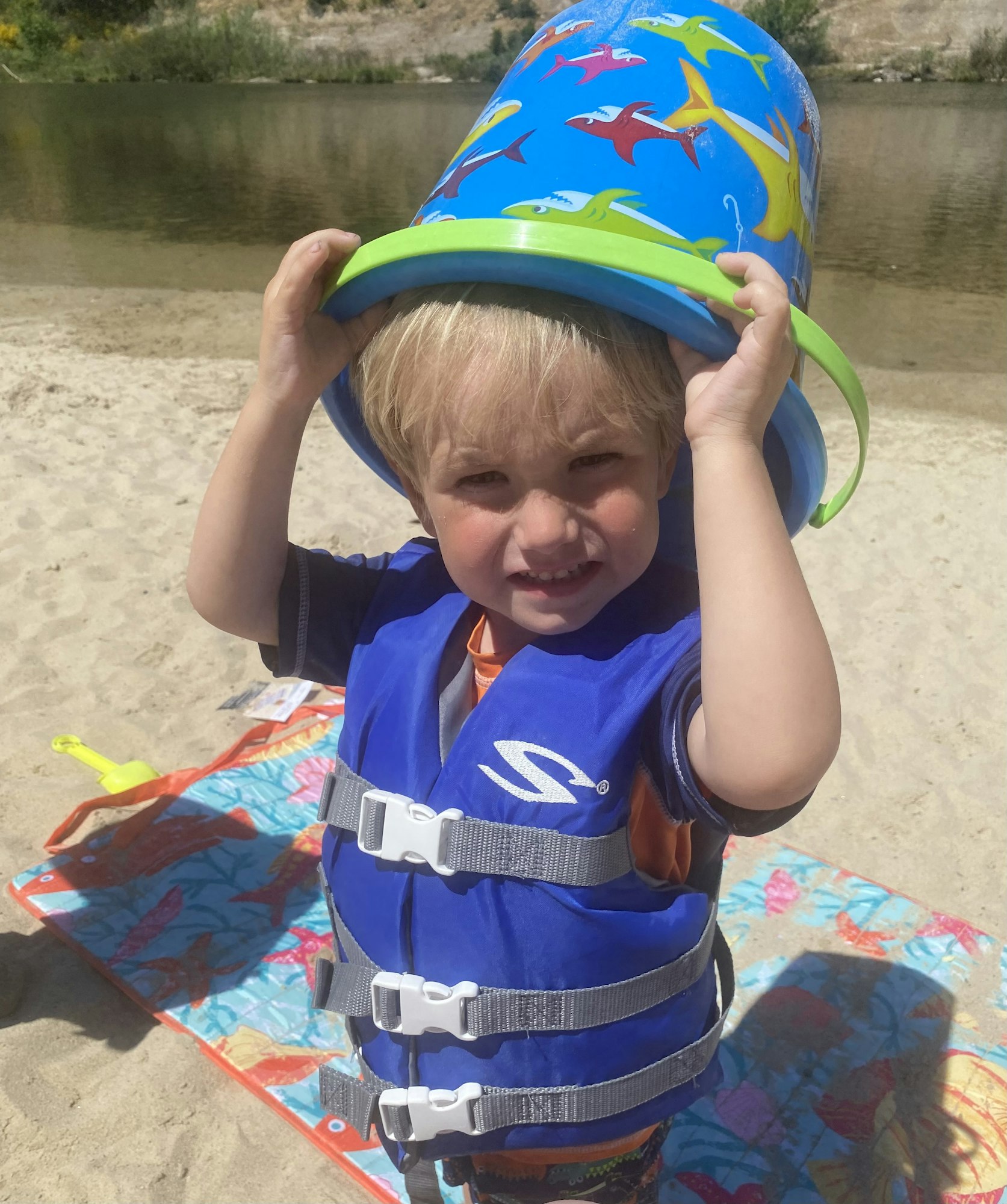 Child on beach wearing life jacket, holding a bucket over head, standing on a towel by the water.