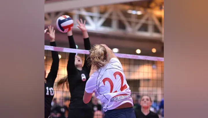 A volleyball game in action, with a player in a white jersey (#22) making a hit while opponents attempt to block.
