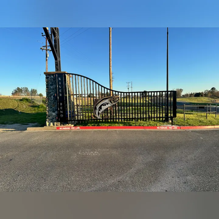 An ornate gate with "RIVERBEND" on it, a fish design, and a "NO PARKING FIRE LANE" sign, under a clear sky.