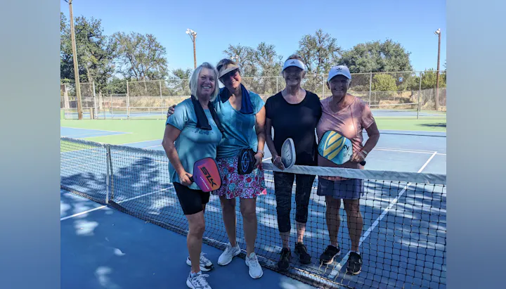 Four women stand together on a tennis court, holding paddles and smiling, with a sunny sky and trees in the background.