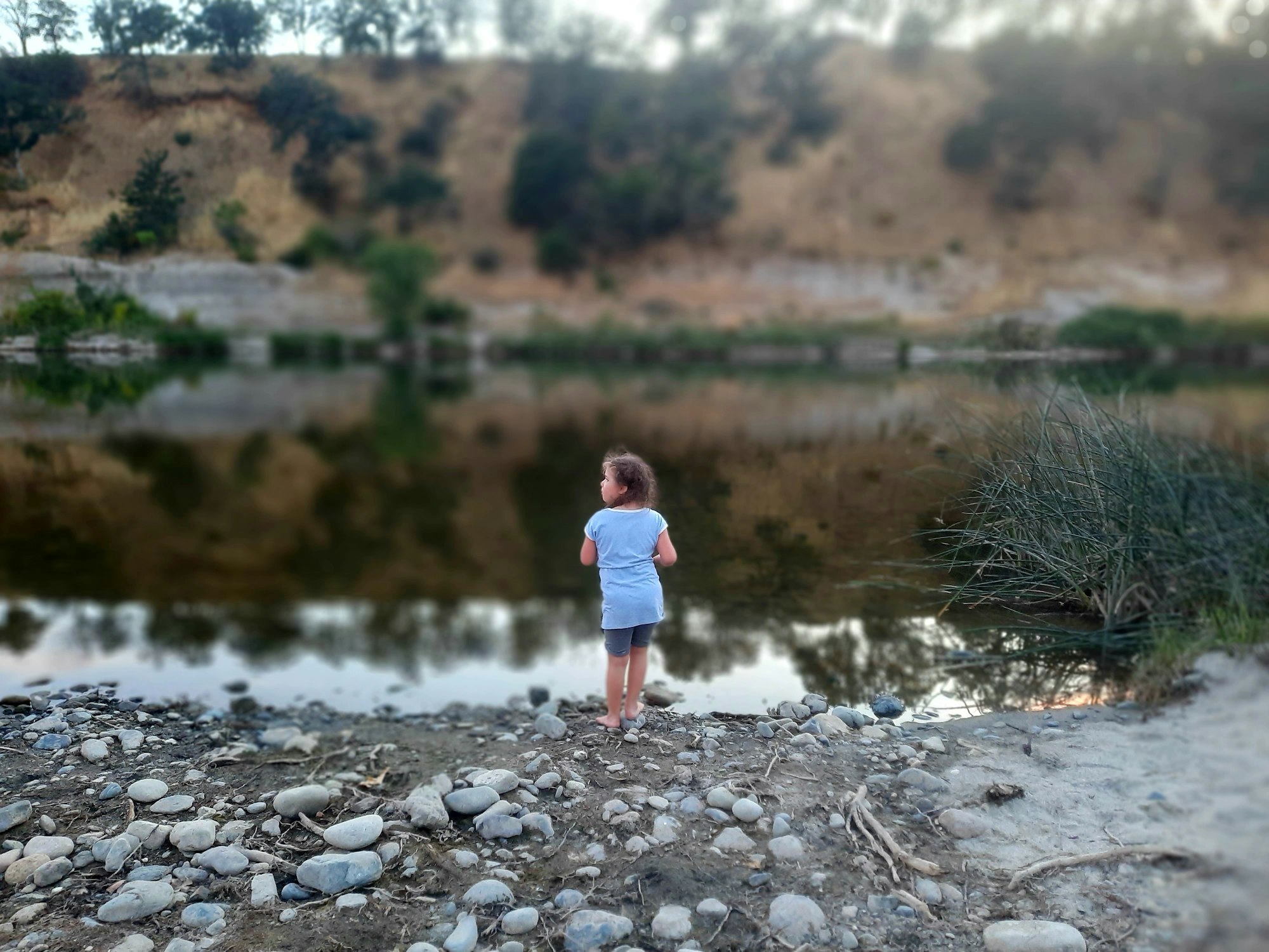 A child stands by a rocky riverside, looking at the calm water with blurred hills in the background.