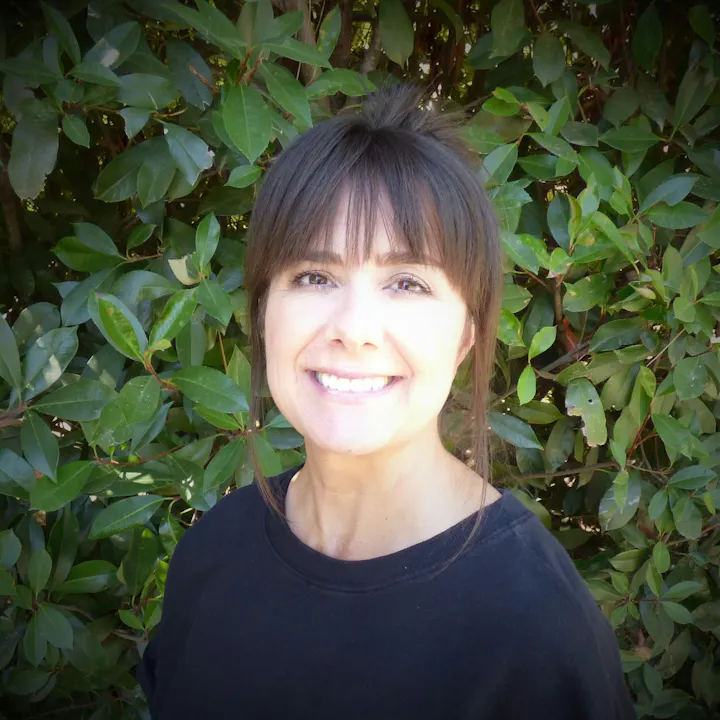 A smiling woman with dark hair, wearing a black shirt, stands in front of a green leafy background.