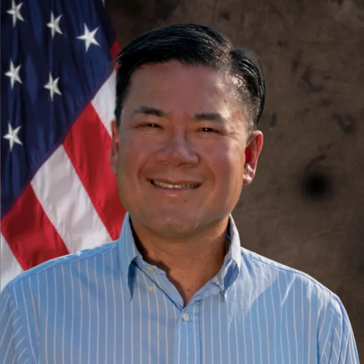 A person smiling, wearing a blue shirt, in front of a U.S. flag.