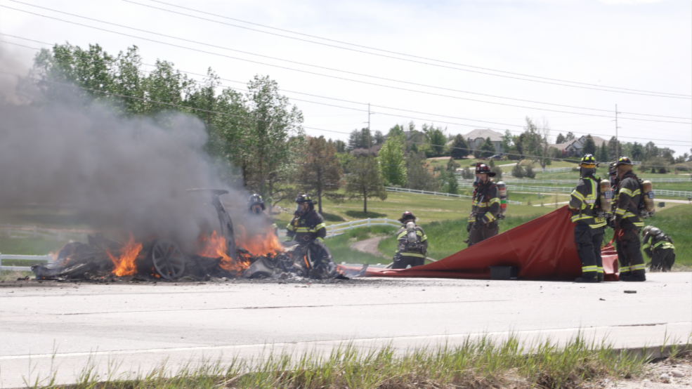 Firefighters extinguishing a burning car on a road.