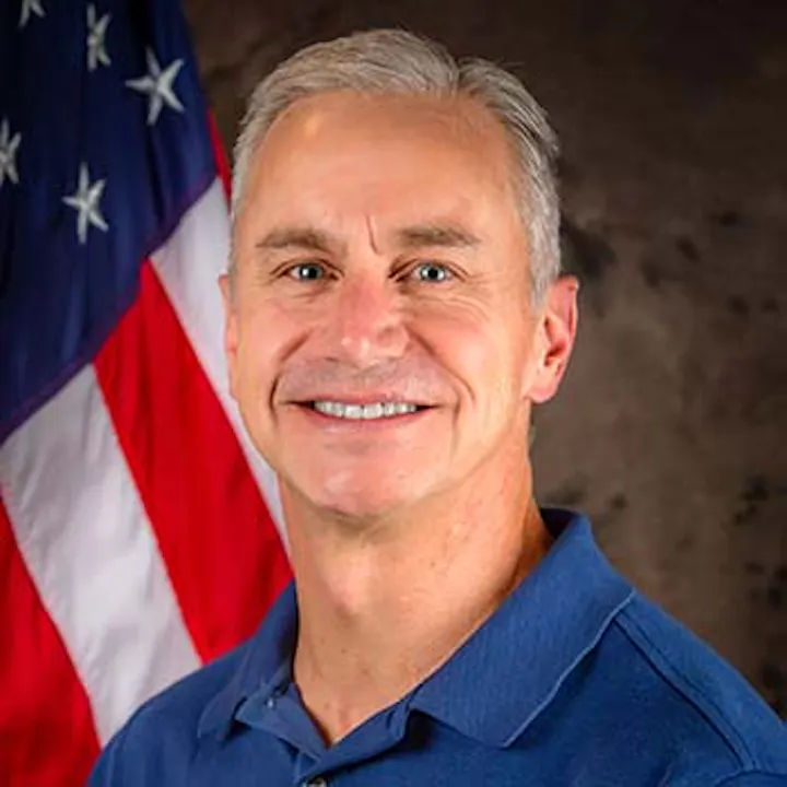 A person smiling in front of a U.S. flag, wearing a blue polo shirt.