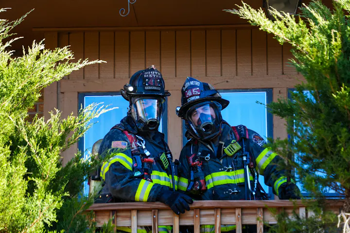 Two firefighters in full gear pose on a balcony, surrounded by greenery, ready for action.