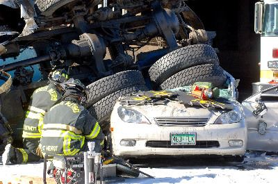 Firefighters are attending to a car crushed under a large vehicle with multiple tires, possibly a truck, in a snow-covered area.