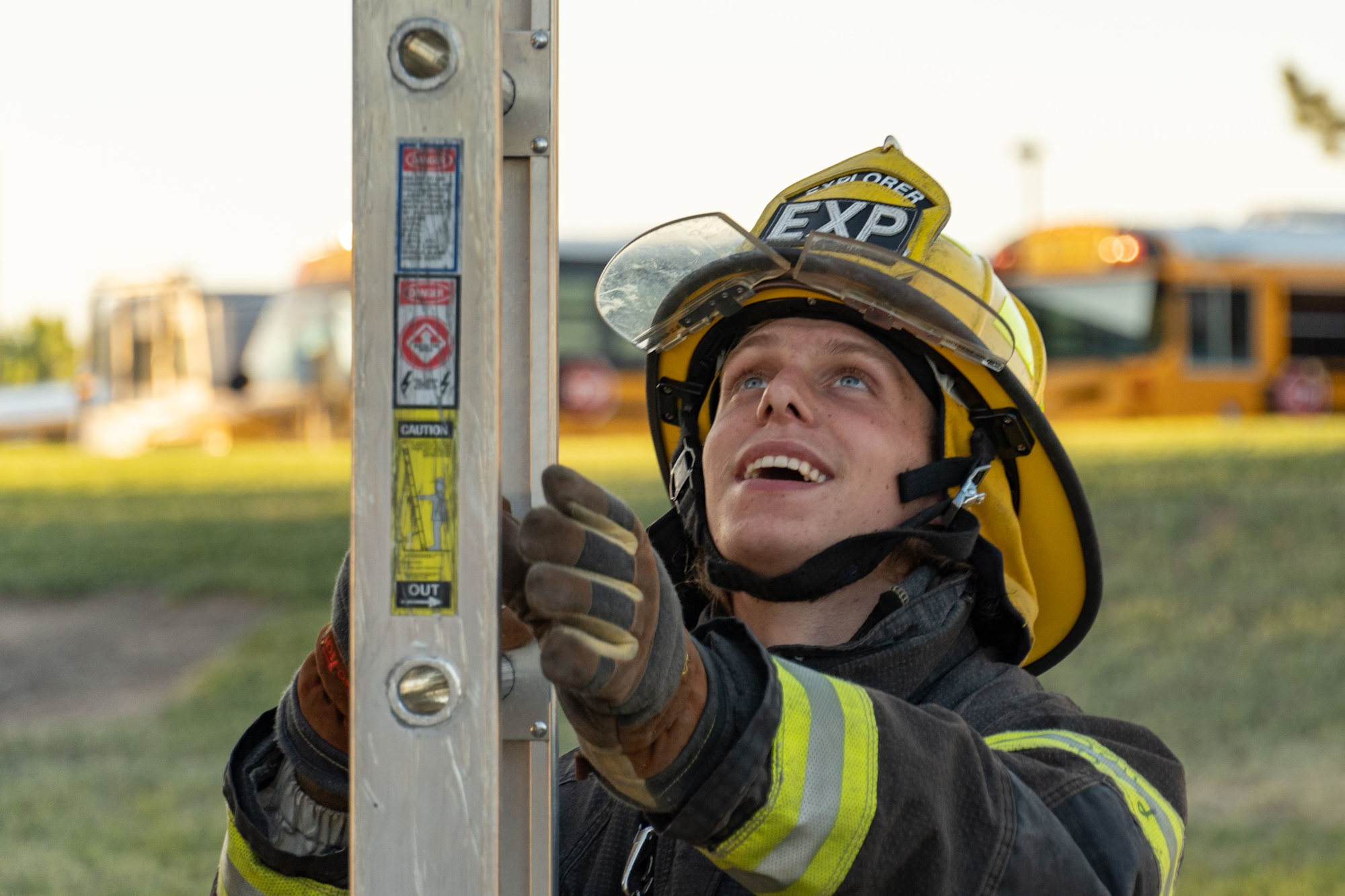 A firefighter in gear is smiling while working with a ladder, set against a sunny outdoor background with school buses nearby.