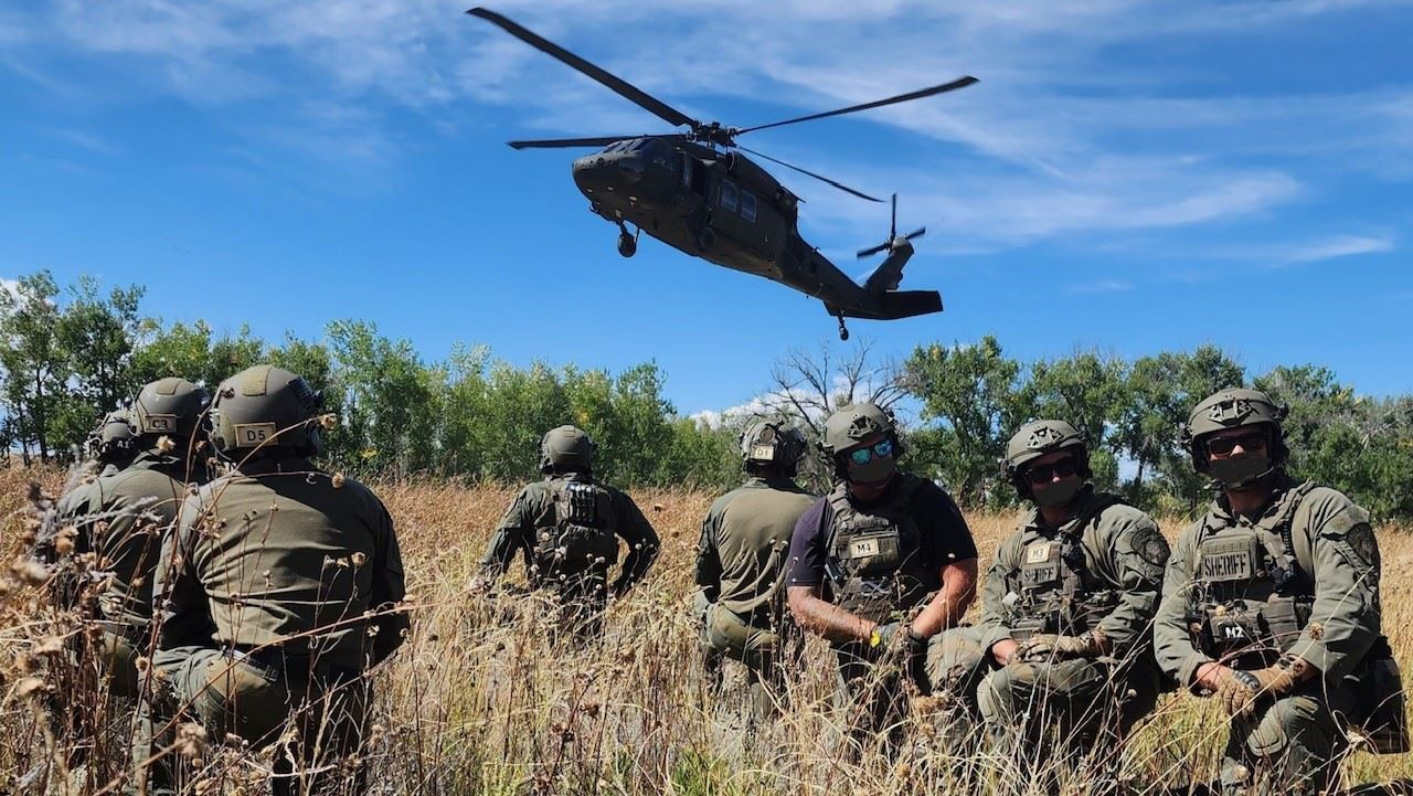 Military personnel in tactical gear and helmets in a field with a helicopter hovering above.