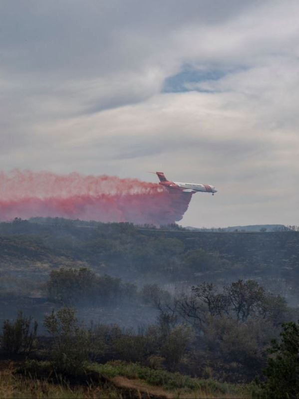 A firefighting plane releases red fire retardant over a smoky landscape, with clouds in the sky.