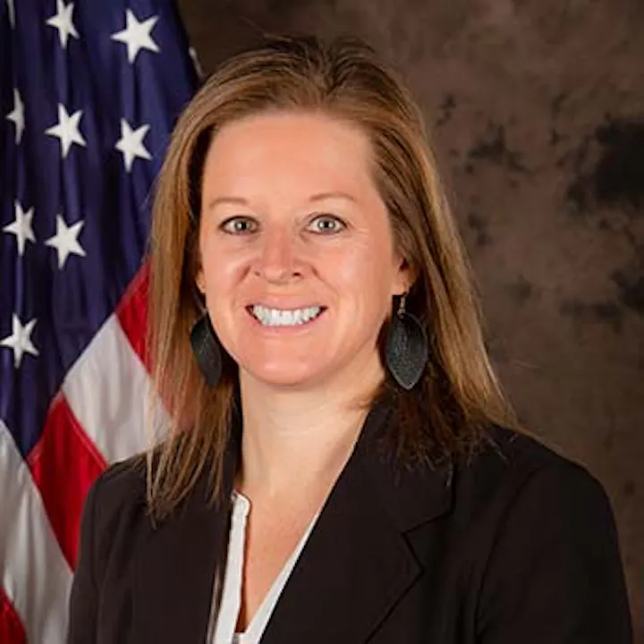 The image shows a smiling woman in front of an American flag, wearing a black blazer and earrings.
