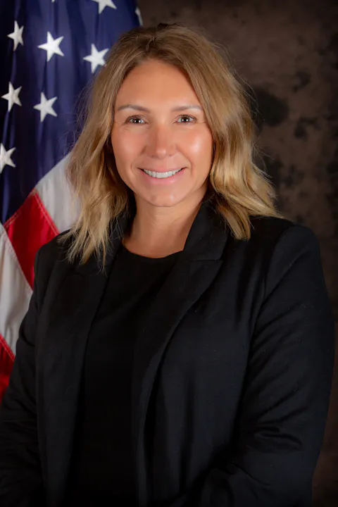 A person in formal attire stands in front of an American flag.