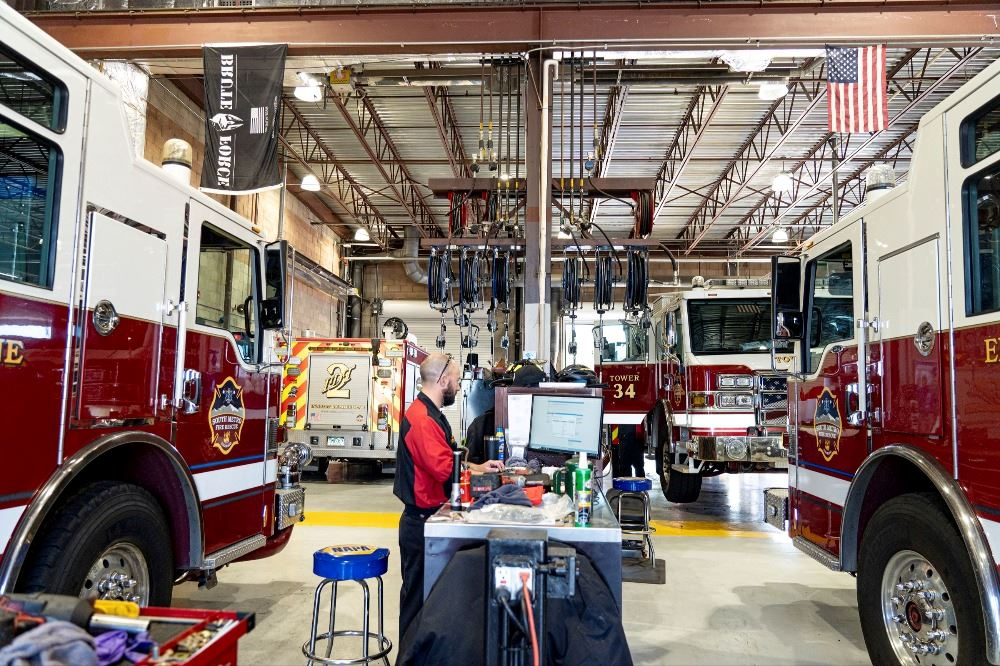 Fire station garage with multiple fire trucks, a technician working at a computer, and flags hanging from the ceiling.