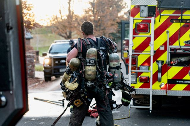 A firefighter carries equipment near a fire truck on a street, with emergency vehicles in the background.