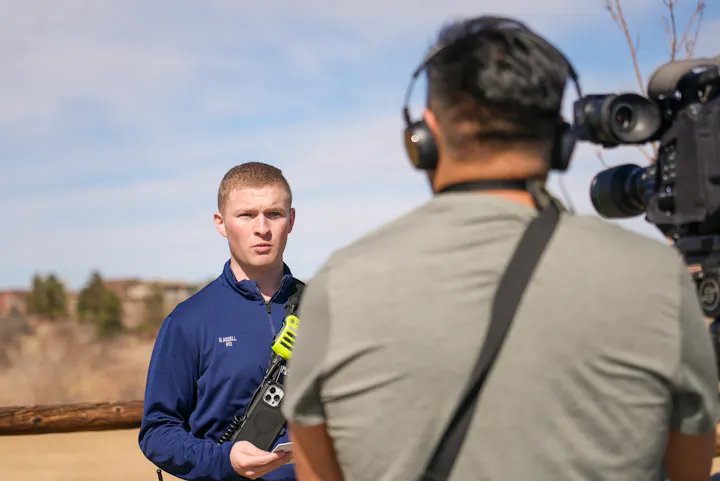 A person in a blue shirt holds a device while being interviewed by a cameraman in an outdoor setting.