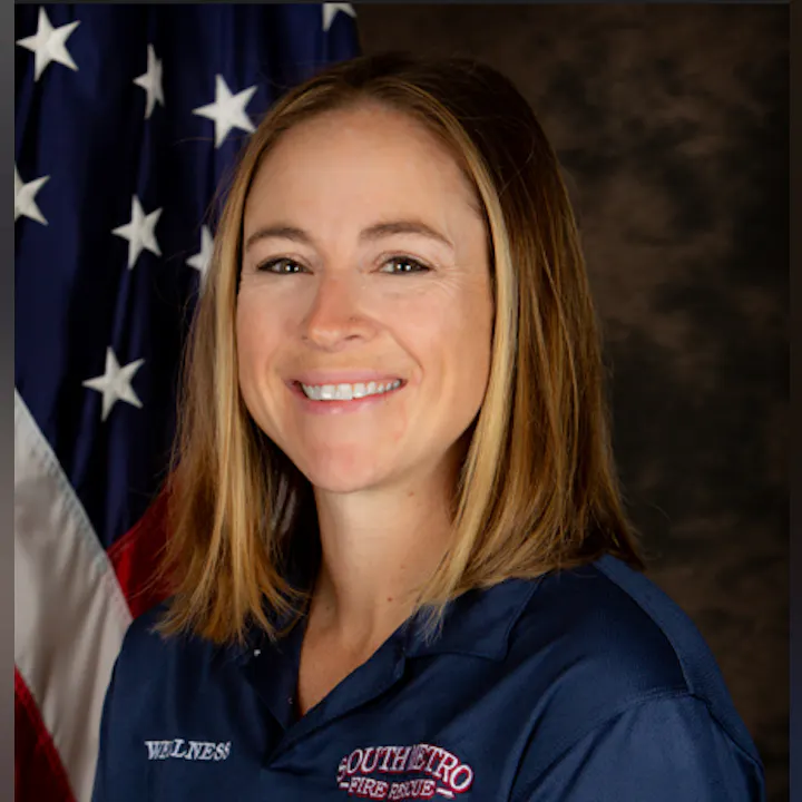 The image shows a smiling woman in a blue shirt with "SOUTHEASTRO FIRE RESCUE" on it, standing in front of an American flag.