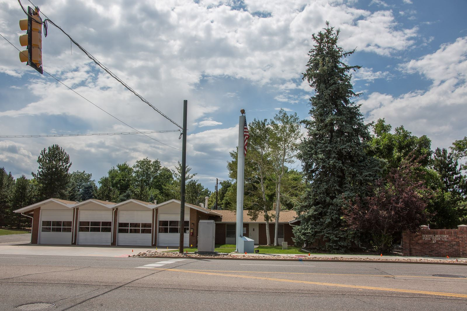 A fire station with three garage doors, a flagpole, large trees, and a traffic light in the foreground.