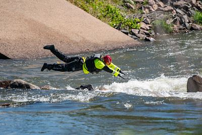 A person in a neon jacket and helmet is diving into a flowing river, likely for a rescue or training exercise.