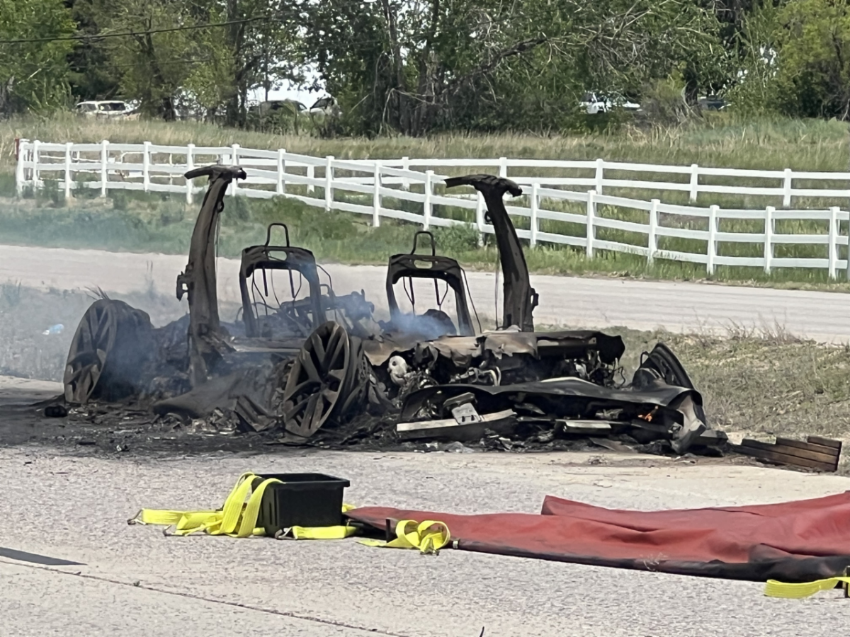 Charred remains of a vehicle with burnt frame and wheels on the road; red barriers and yellow straps in the foreground.