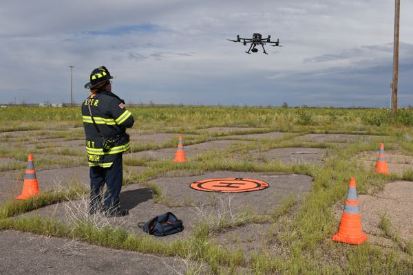 Firefighter operates a drone near an orange landing pad on an overgrown field, surrounded by traffic cones.