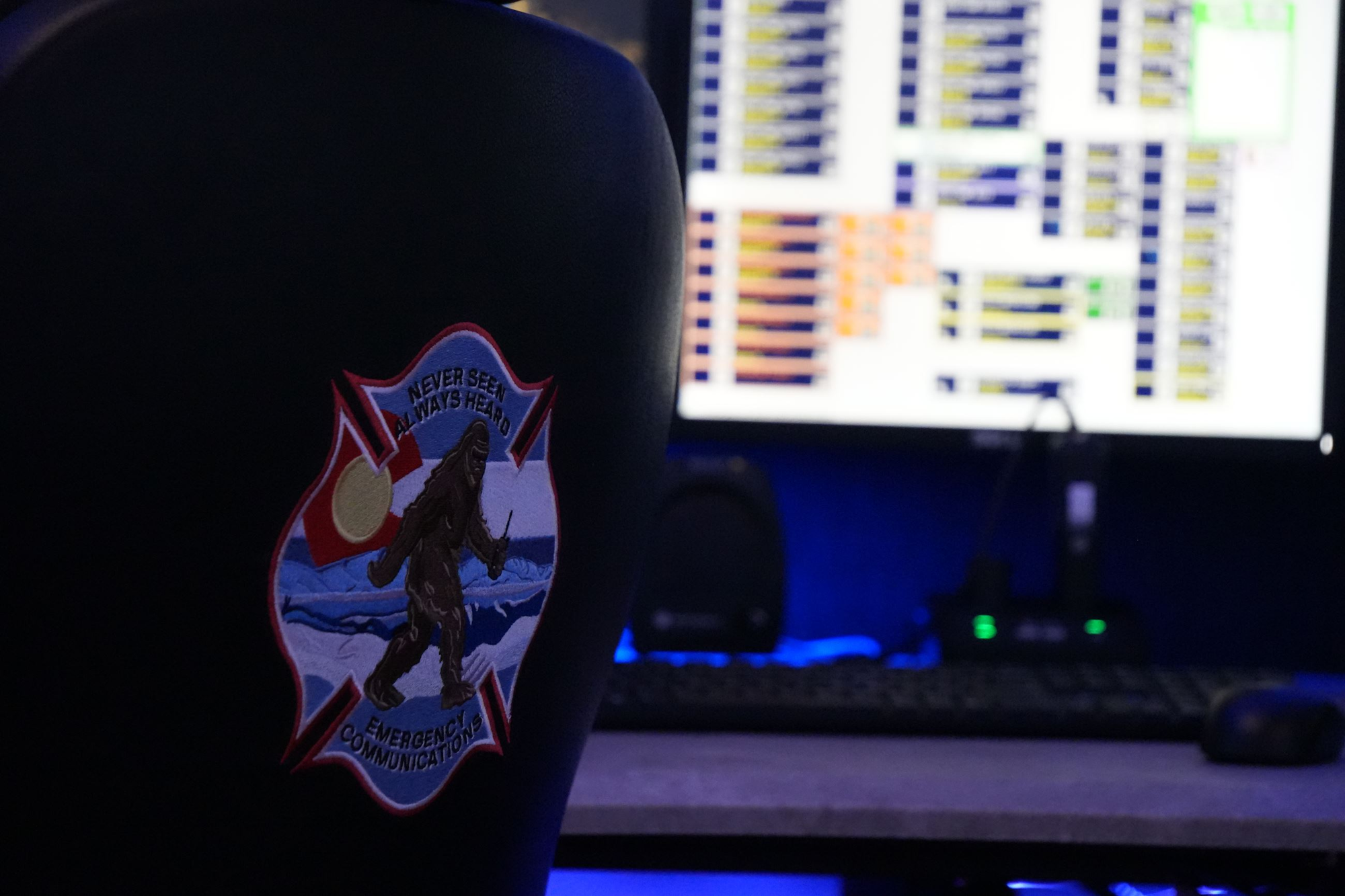 An office setting featuring a chair with a patch depicting a Bigfoot-like creature and a screen displaying data or charts.