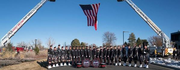 A group of people in uniform with drums, standing under a large American flag hanging between two fire truck ladders.