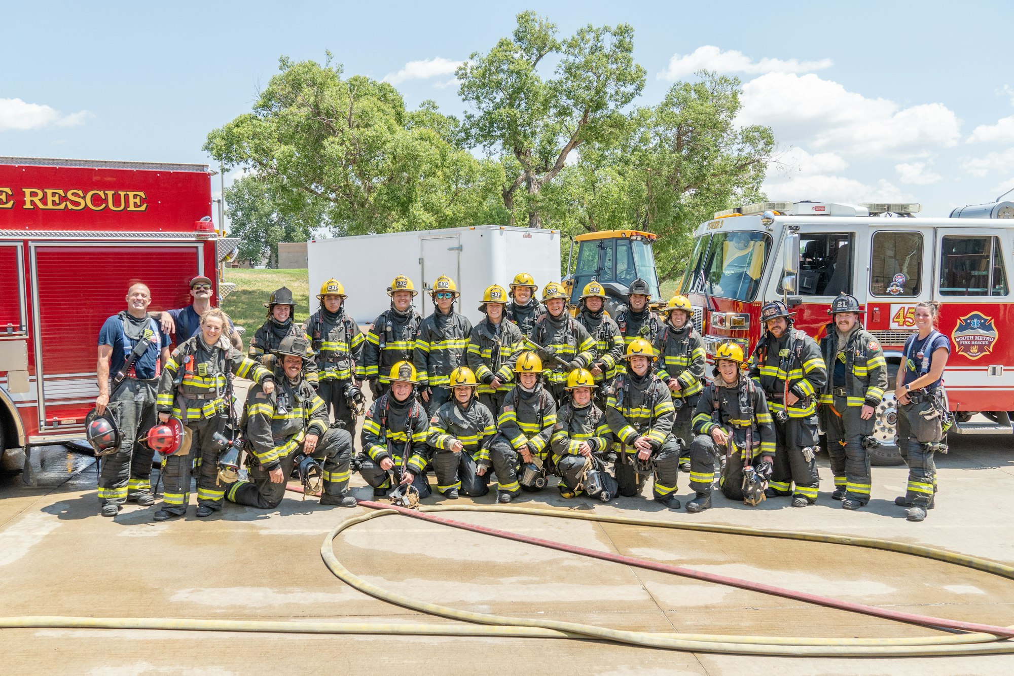 A group photo of firefighters in uniform, posing in front of fire trucks and equipment on a sunny day.