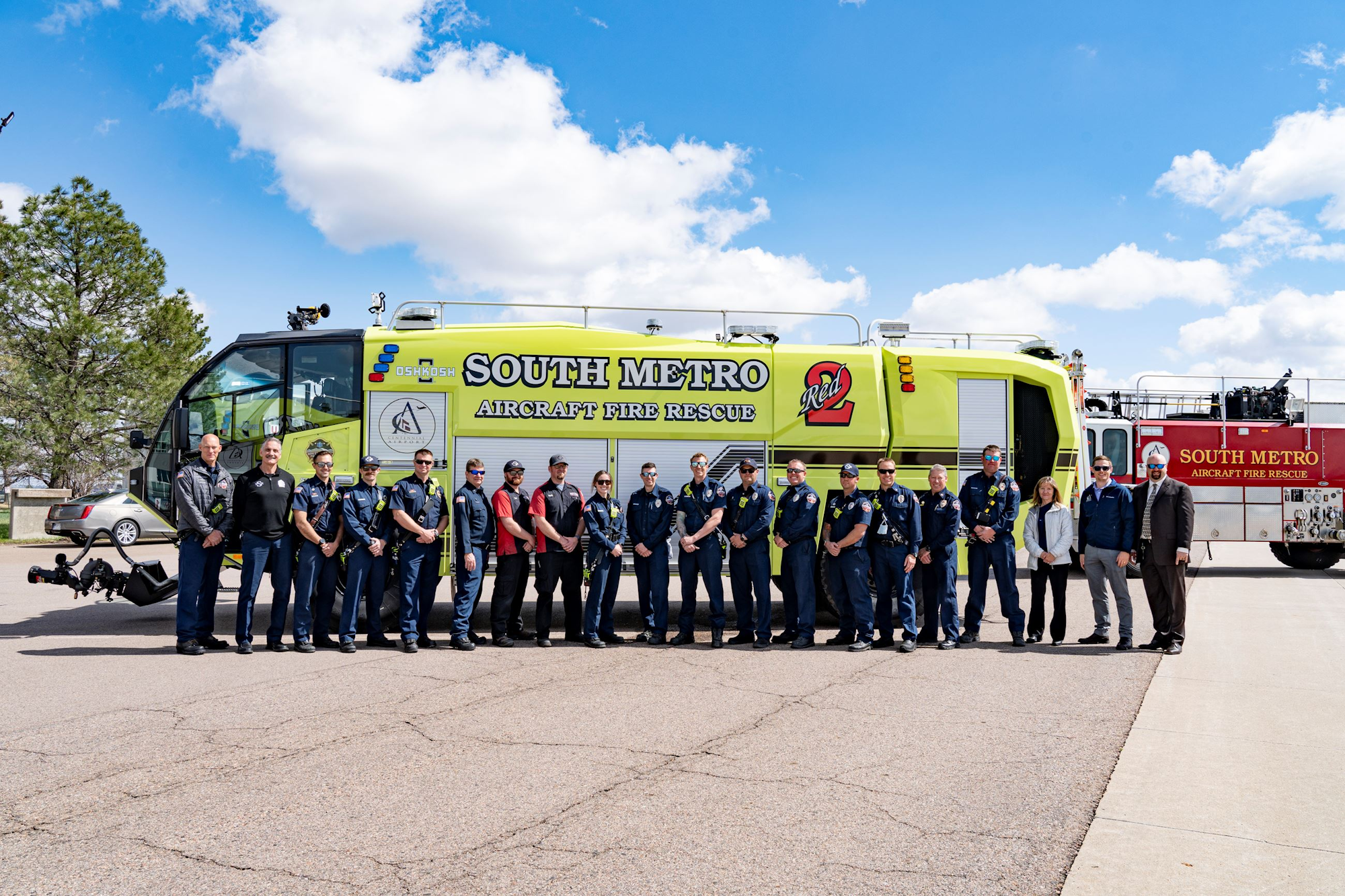 A group of uniformed firefighters stands beside a "South Metro Aircraft Fire Rescue" vehicle under a blue sky.