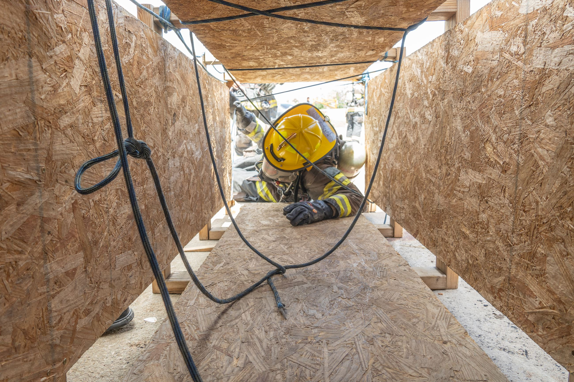 A firefighter in protective gear crawls through a narrow space, focusing on a training exercise amidst wood barriers.