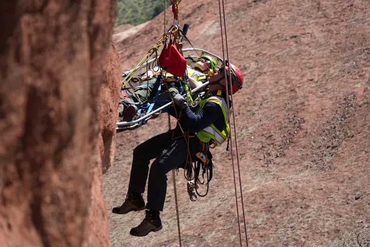 A rescue worker is hoisting a person on a stretcher up a rocky cliff, using ropes and safety gear in a mountainous area.