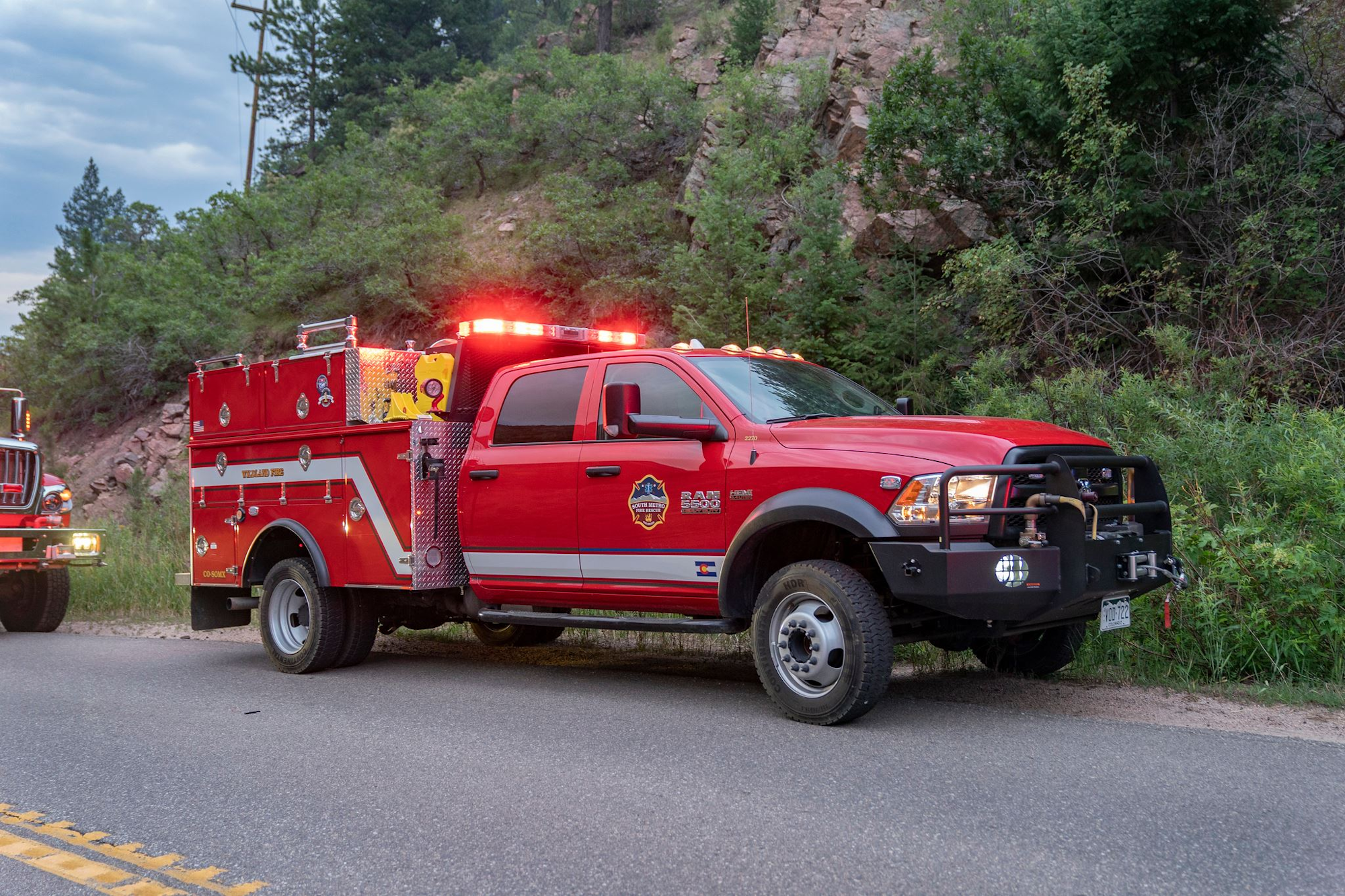 A red fire truck with flashing lights is parked on a road beside a wooded area.