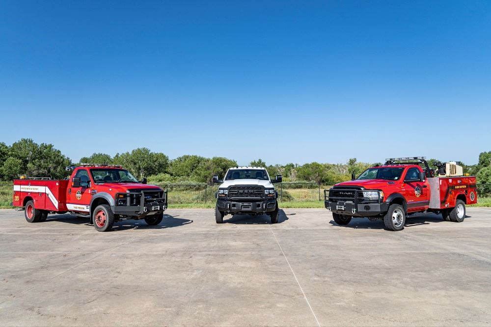 Three trucks parked on a concrete surface, two red and one white, with a clear blue sky and trees in the background.