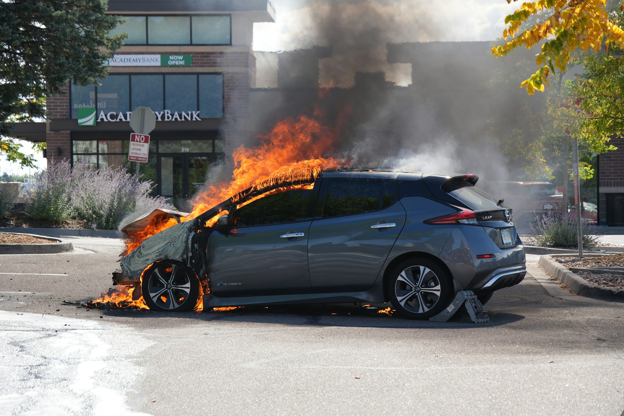 A car is on fire in a parking lot, with visible flames and smoke.